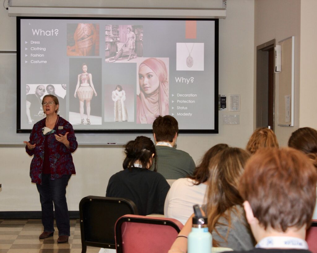 Instructor speaks in the front of a full classroom with a media presentation projected on the screen behind her.