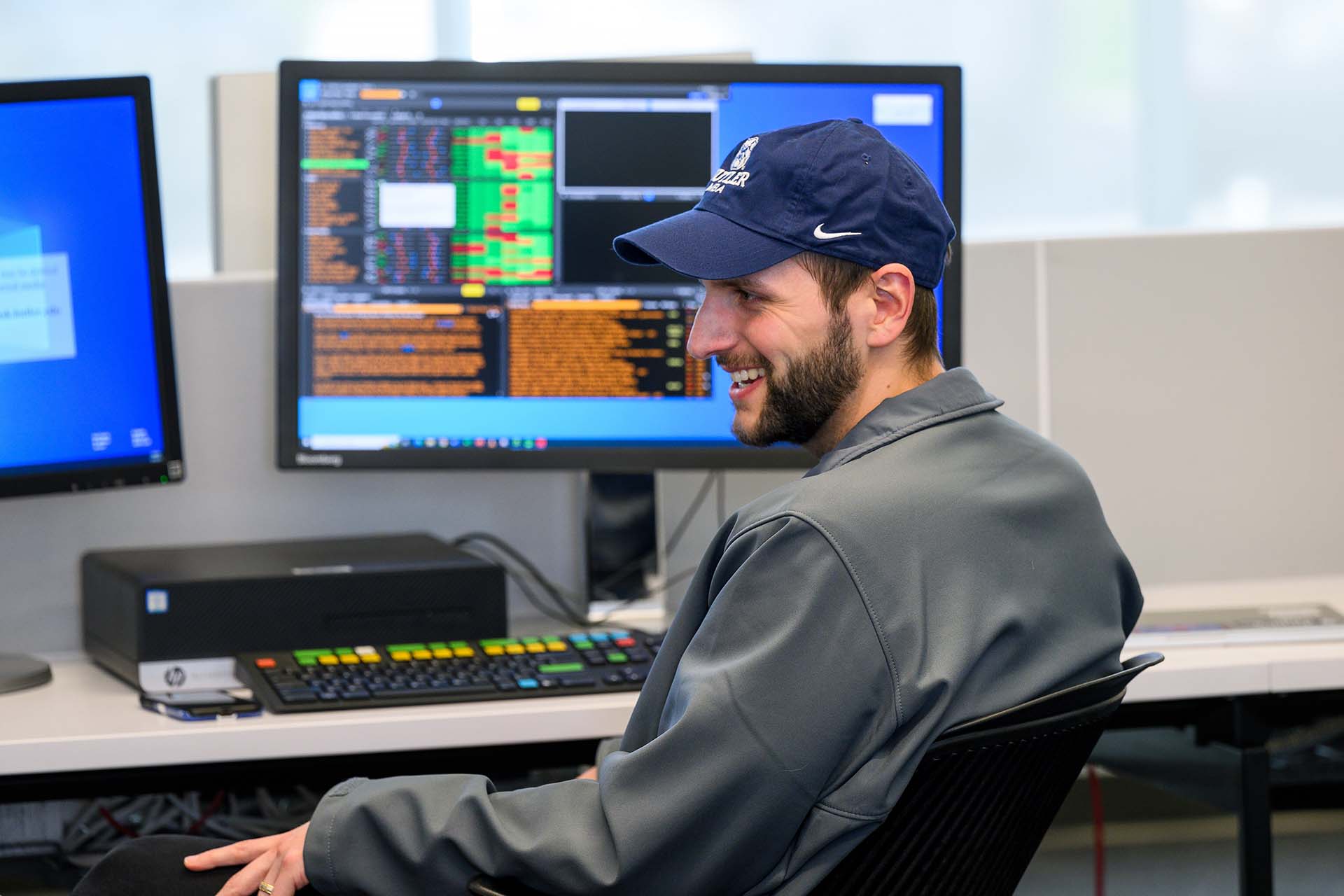Man in Butler hat smiles at desk with computer monitors.