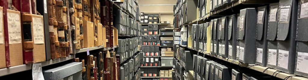 View down an aisle of University Archives shelving, with old ledgers and gray archival boxes filling the shelves.