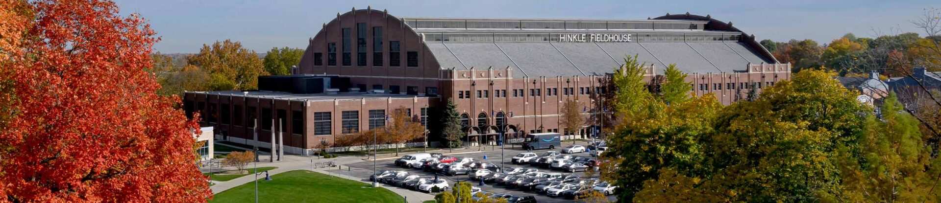 Butler's brick Hinkle Fieldhouse basketball arena on a sunny autumn day. Shade trees in their fall colors and a full parking lot appear in the foreground.