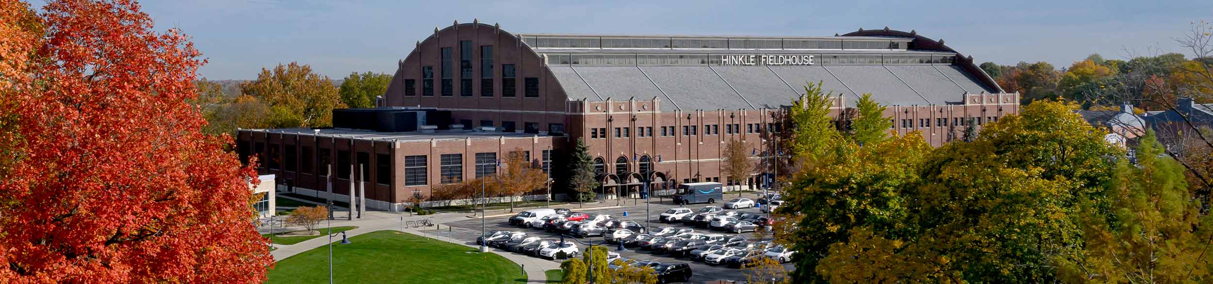 Butler's brick Hinkle Fieldhouse basketball arena on a sunny autumn day. Shade trees in their fall colors and a full parking lot appear in the foreground.