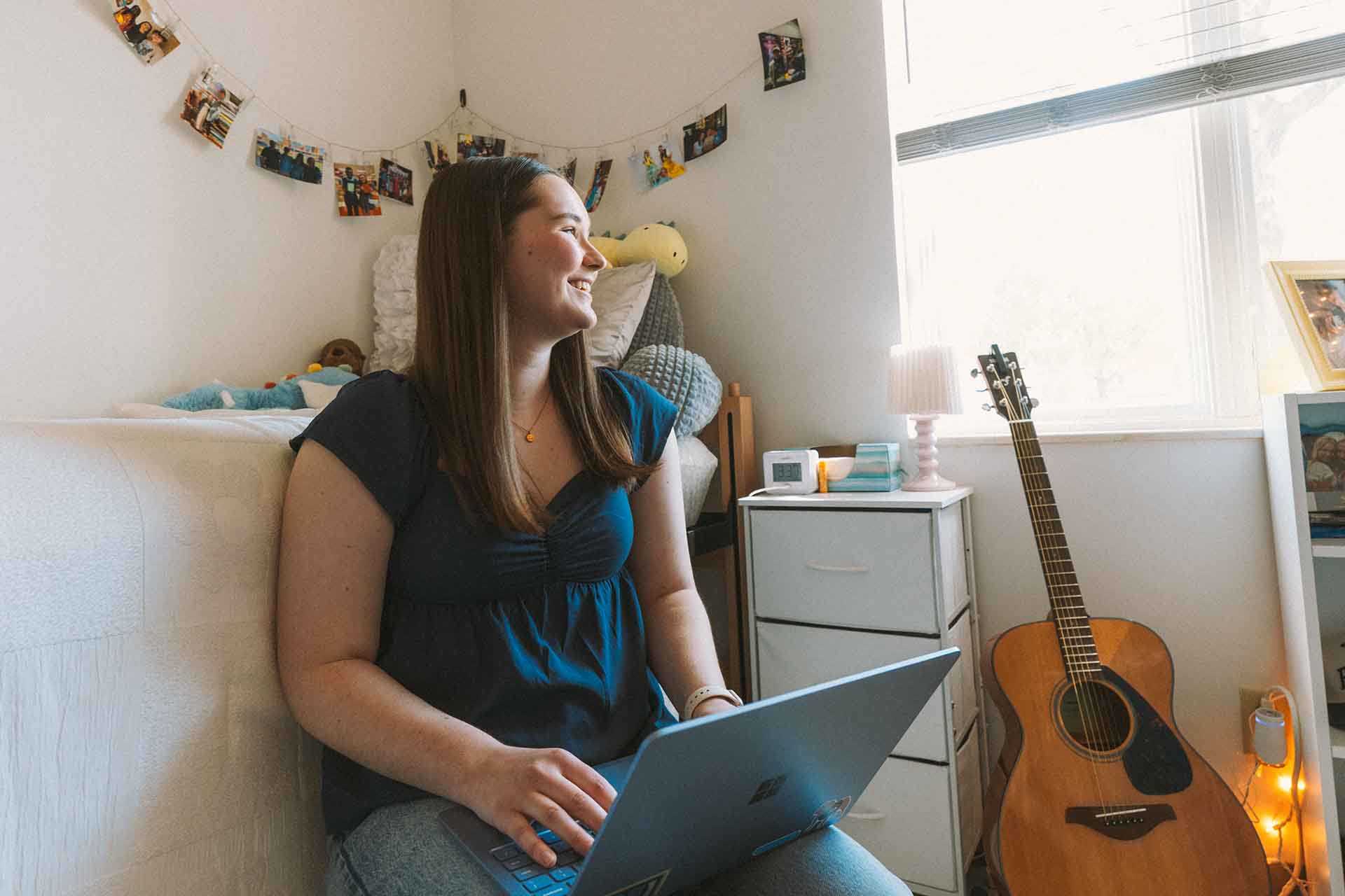 Female college student smiling as she looks out the window of her dorm room.