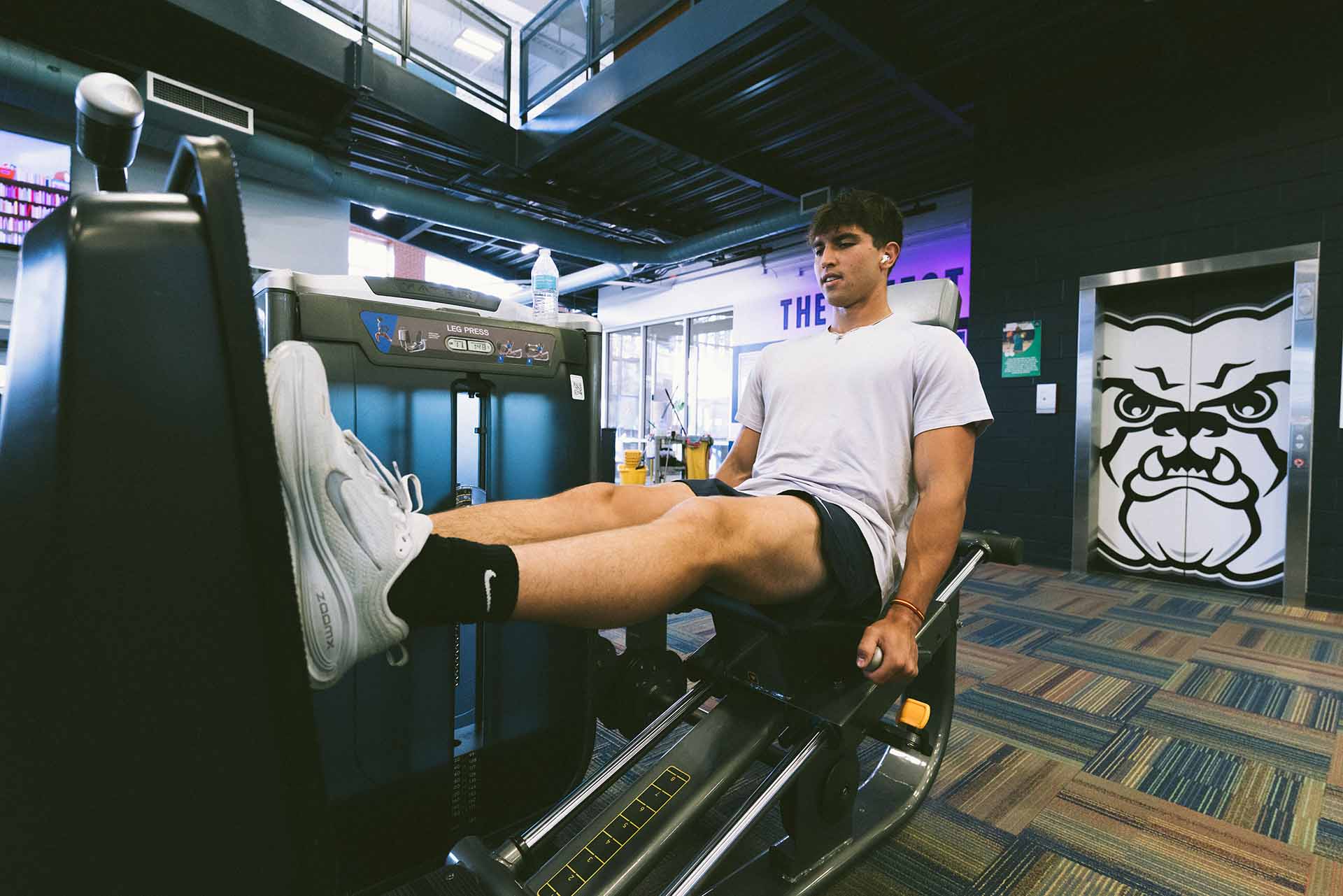 Male college student uses leg press weight machine at campus gym facility.