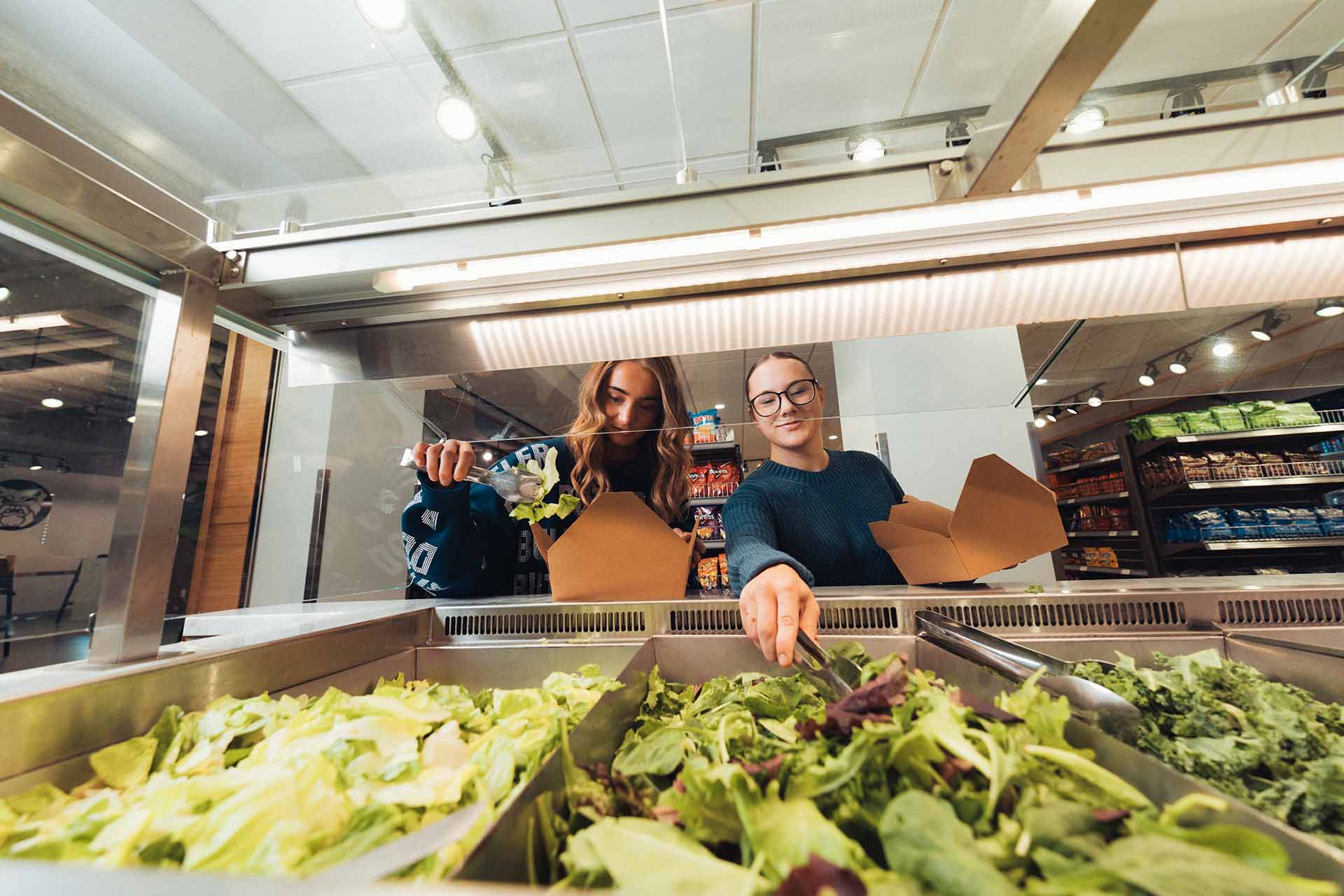 Two college students use tongs to fill their lunch boxes with vegetables from the salad bar.