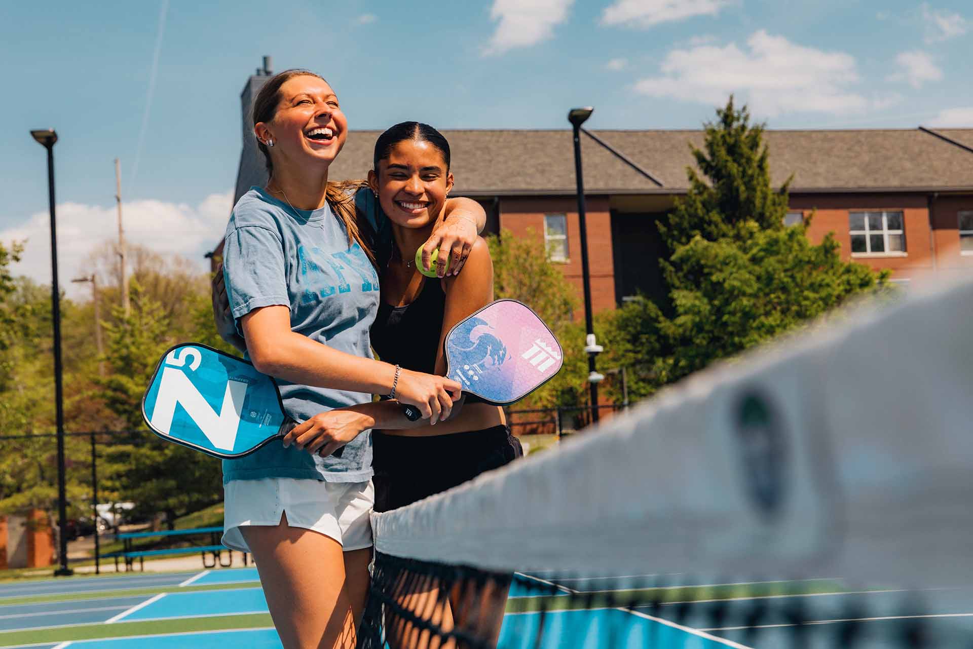 Two female college students laugh and hug at the pickleball court net.