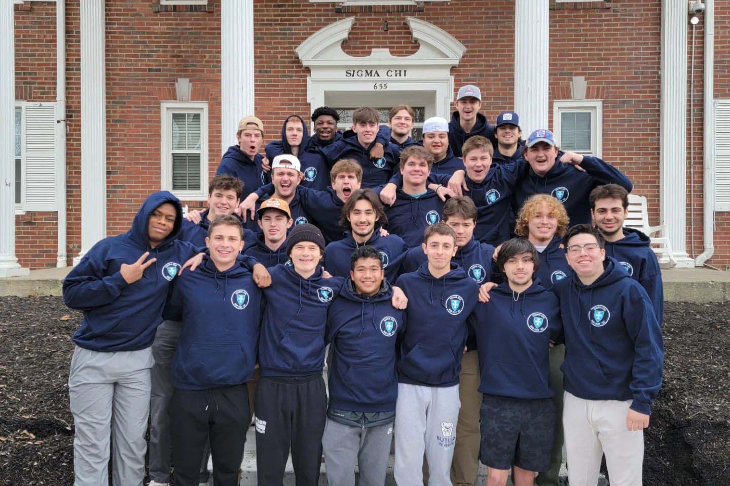 Group of Sigma Chi fraternity brothers wear hoodies and pose for a photo in front of their house.