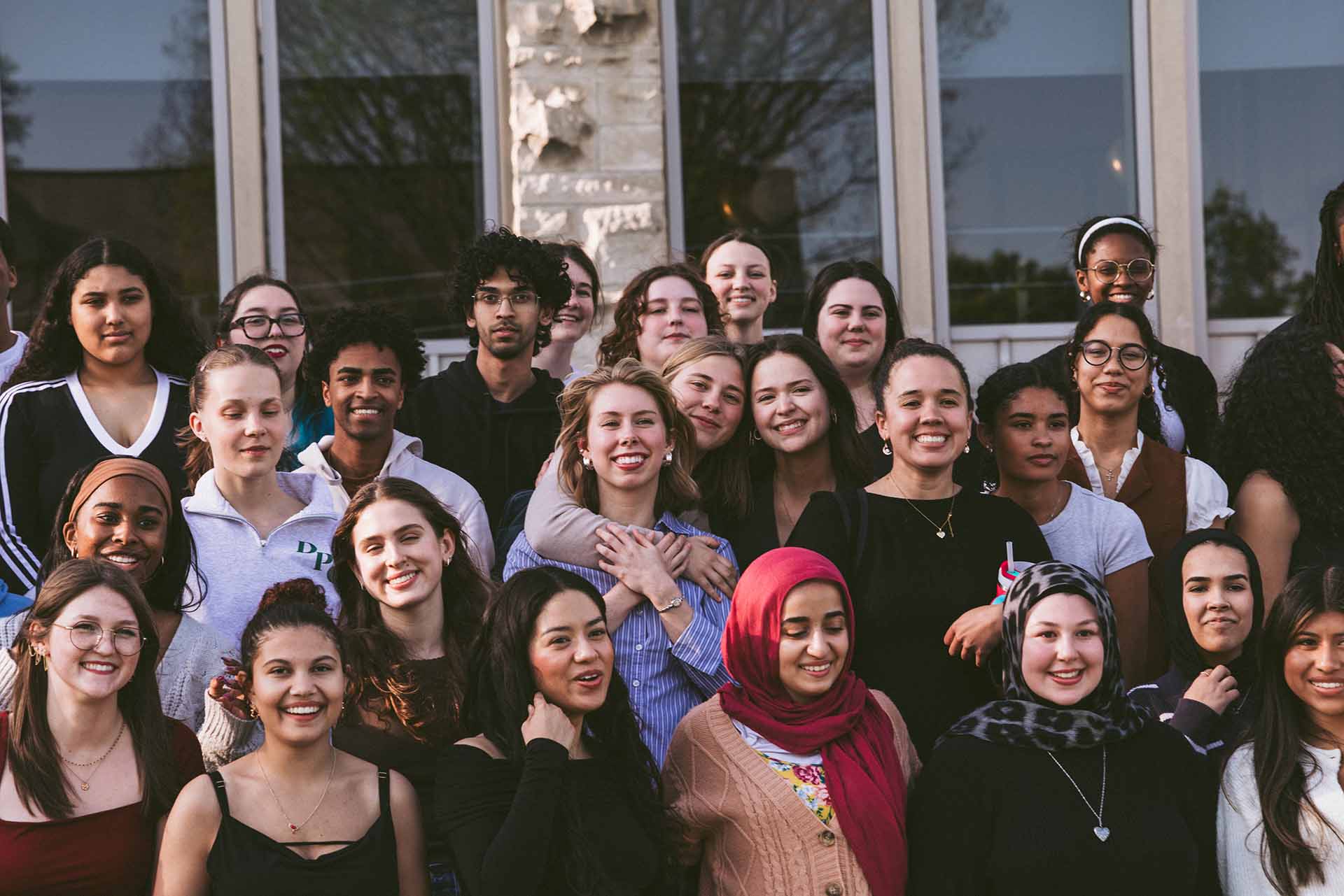 Large group of college students pose for a photo on the steps of a building with large windows behind them.