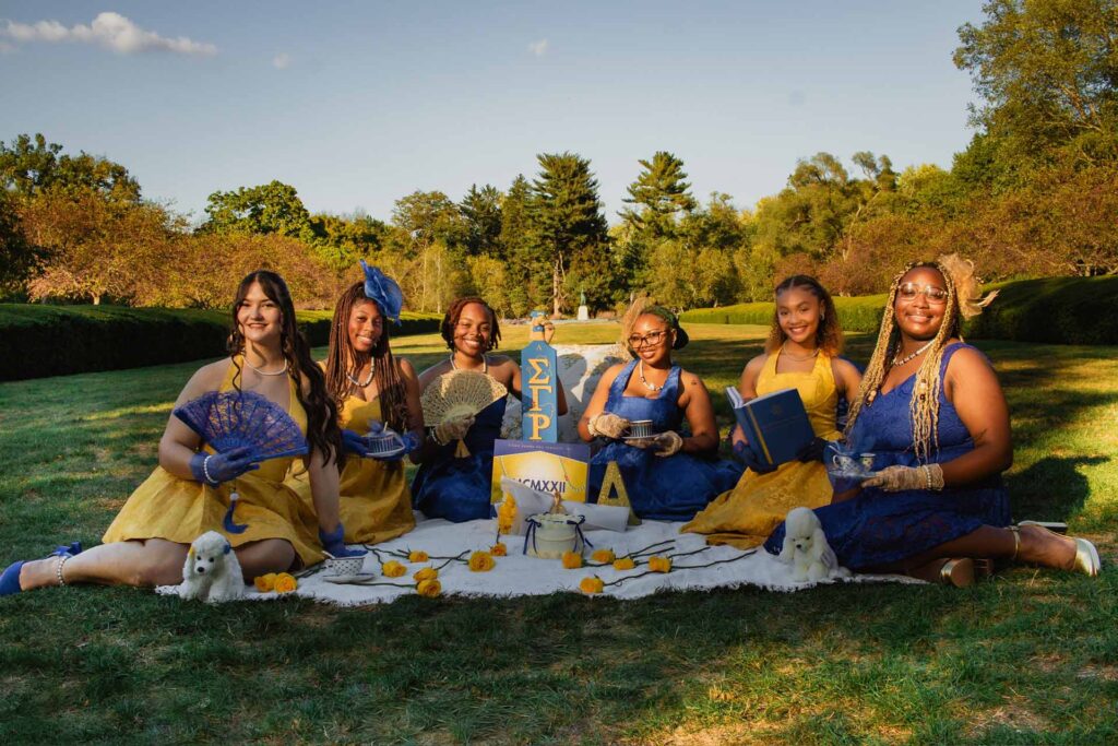 Six women in blue and yellow dresses hold fans and teacups while sitting on a blanket in Holcomb Gardens.