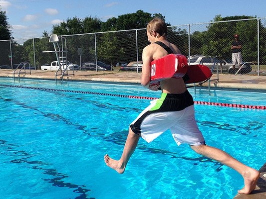 This is a photo of a young guy learning how to be a lifeguard. He is jumping into the pool holding a red rescue tube.