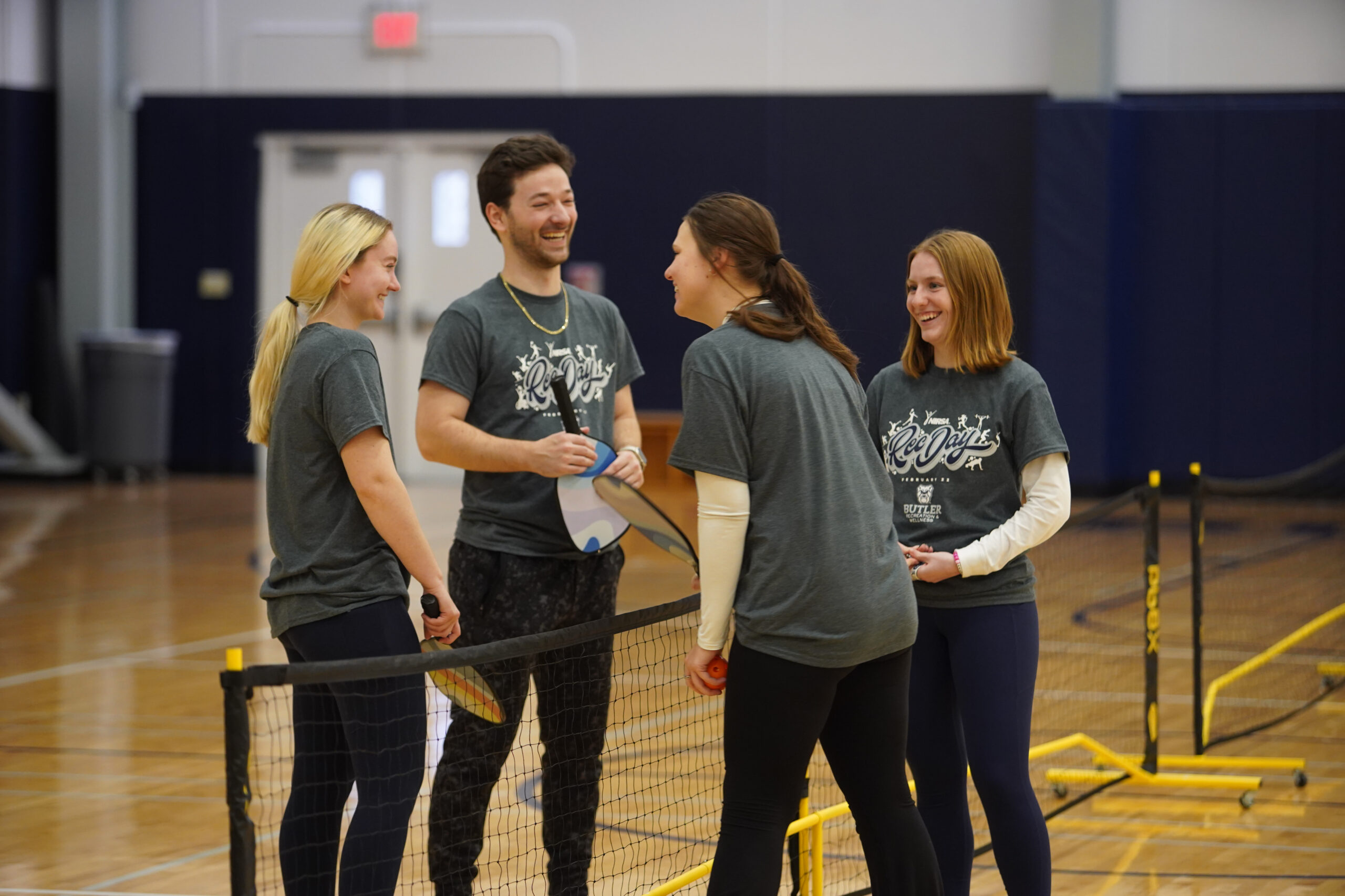This is a photo of four people standing on a basketball court. They are laughing and smiling and look like they are finishing a game of pickleball. 