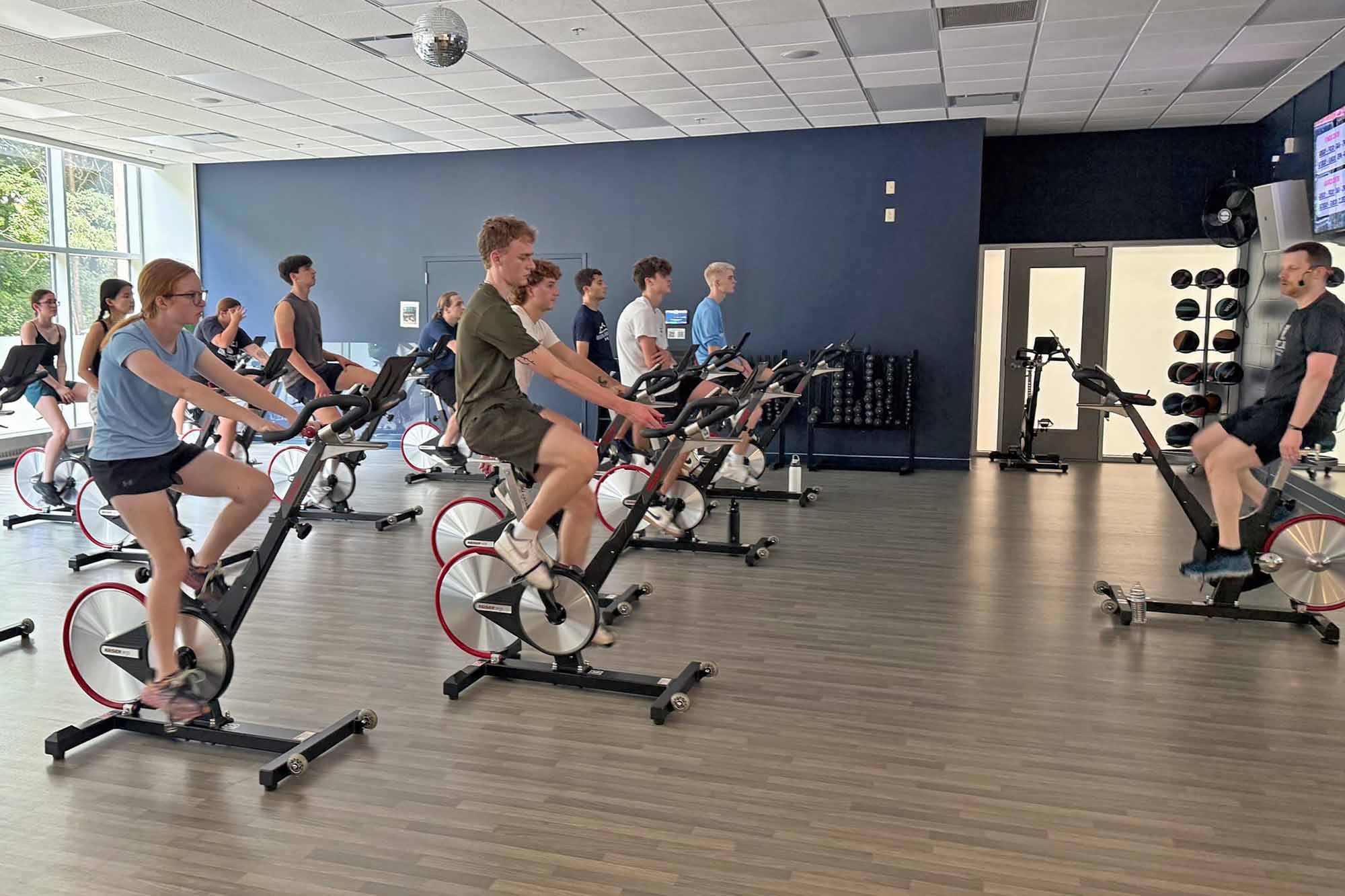 Many students on stationary bikes during cycling class as instructor in front of room guides them.