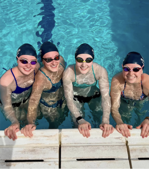 This is a photo of four females in the swimming pool. They are at the edge of the pool, holding onto the side, and smiling up at the camera.