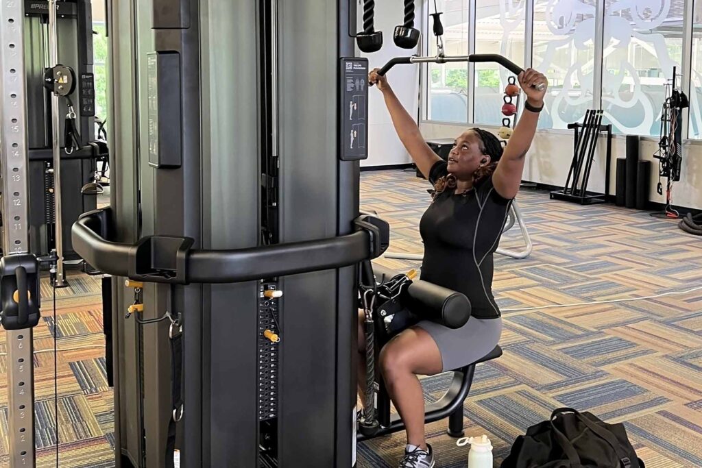 Woman reaches up while using the lat pull-down machine in the Health and Recreation Complex fitness area.