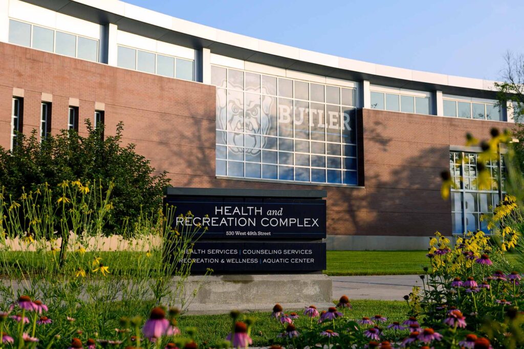 Wildflowers adorn the foreground while the Health and Recreation Complex sign stands in front of the large brick building that has a massive window with the word Butler and bulldog face in it.