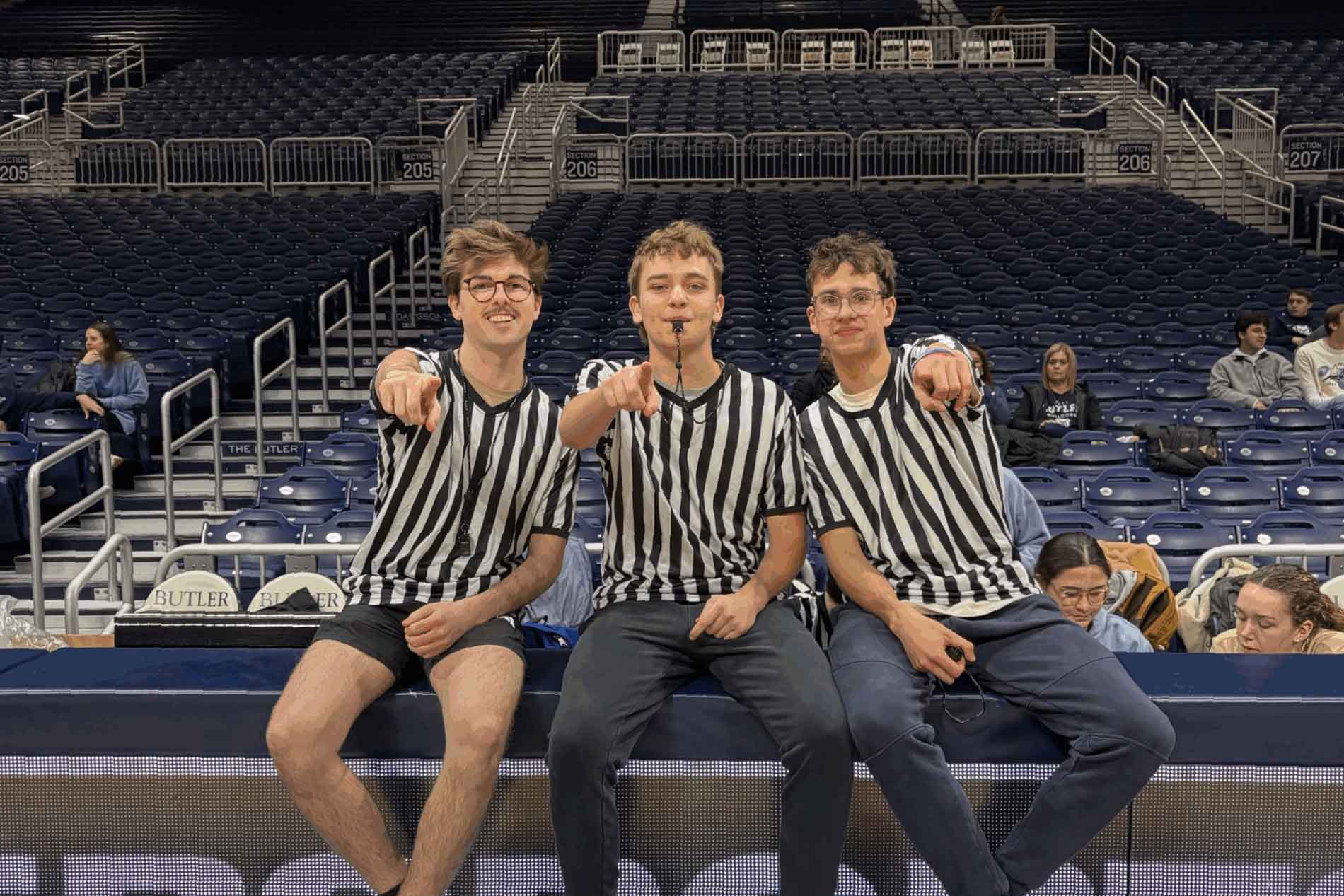 Student referees sit on Hinkle Fieldhouse scorer's table and point toward the camera.