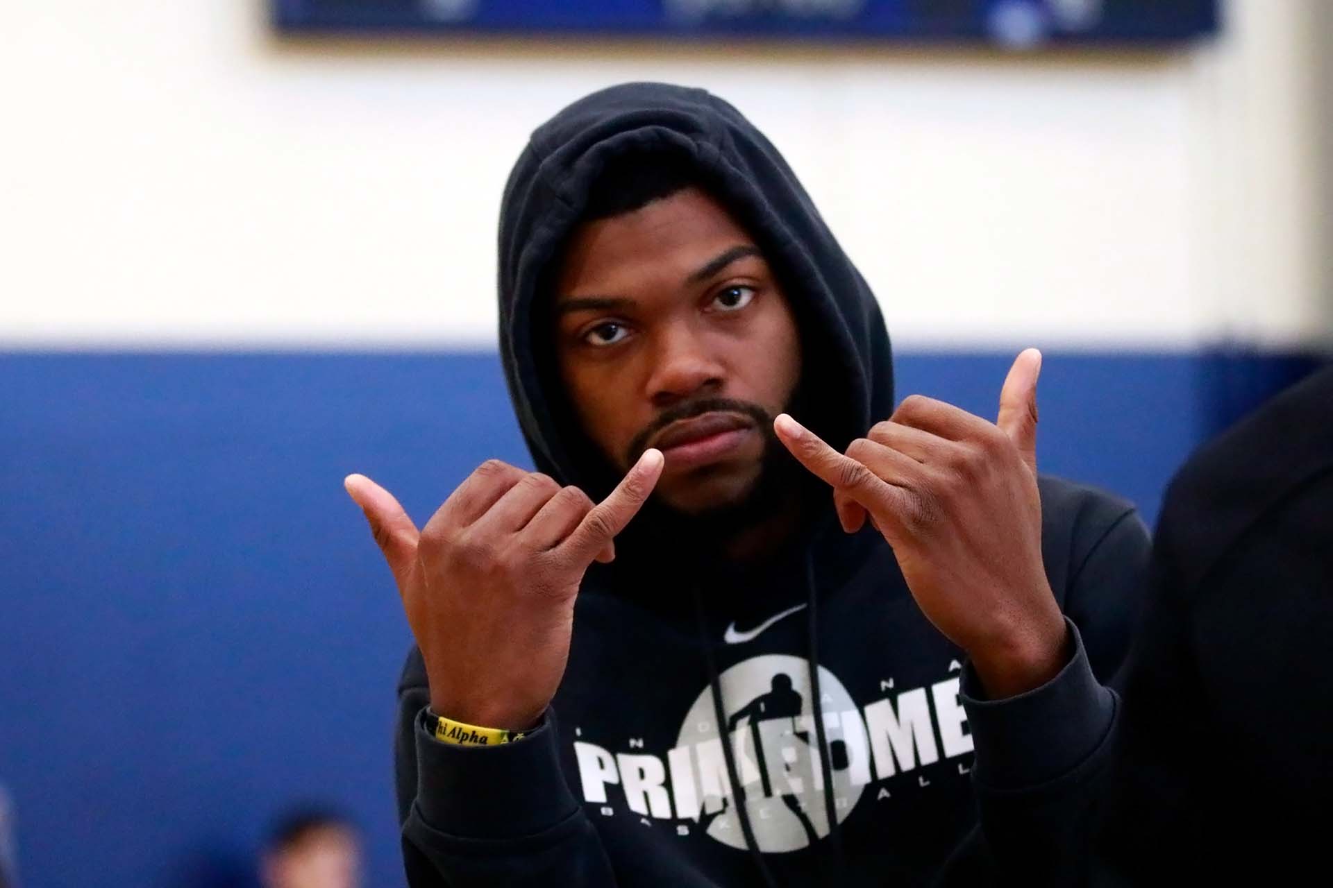 A man on a basketball court wearing a hoodie uses both hands to give the "hang loose" greeting.
