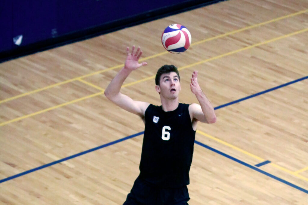 Man wearing an athletic jersey with a Butler bulldog logo winds up as he is serving a volleyball.