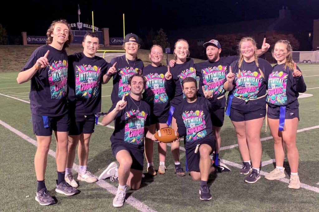 Team of intramural flag football champions smile and make the "Number One" sign as they celebrate on football field.
