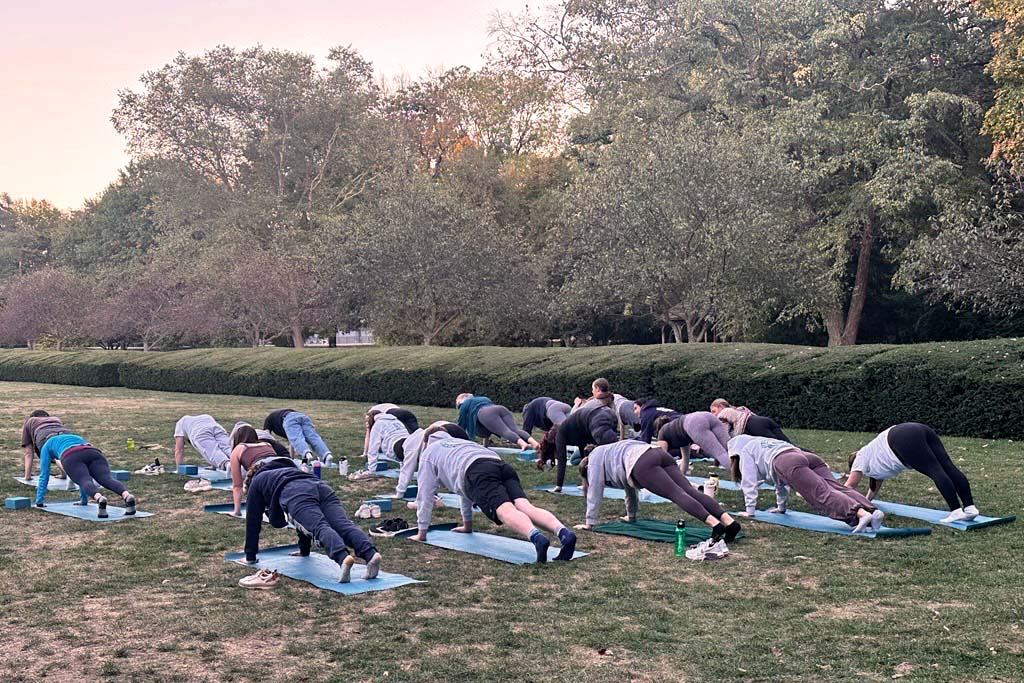 Many people in a fitness class stretching on their mats in the green lawn of the Holcomb Gardens.