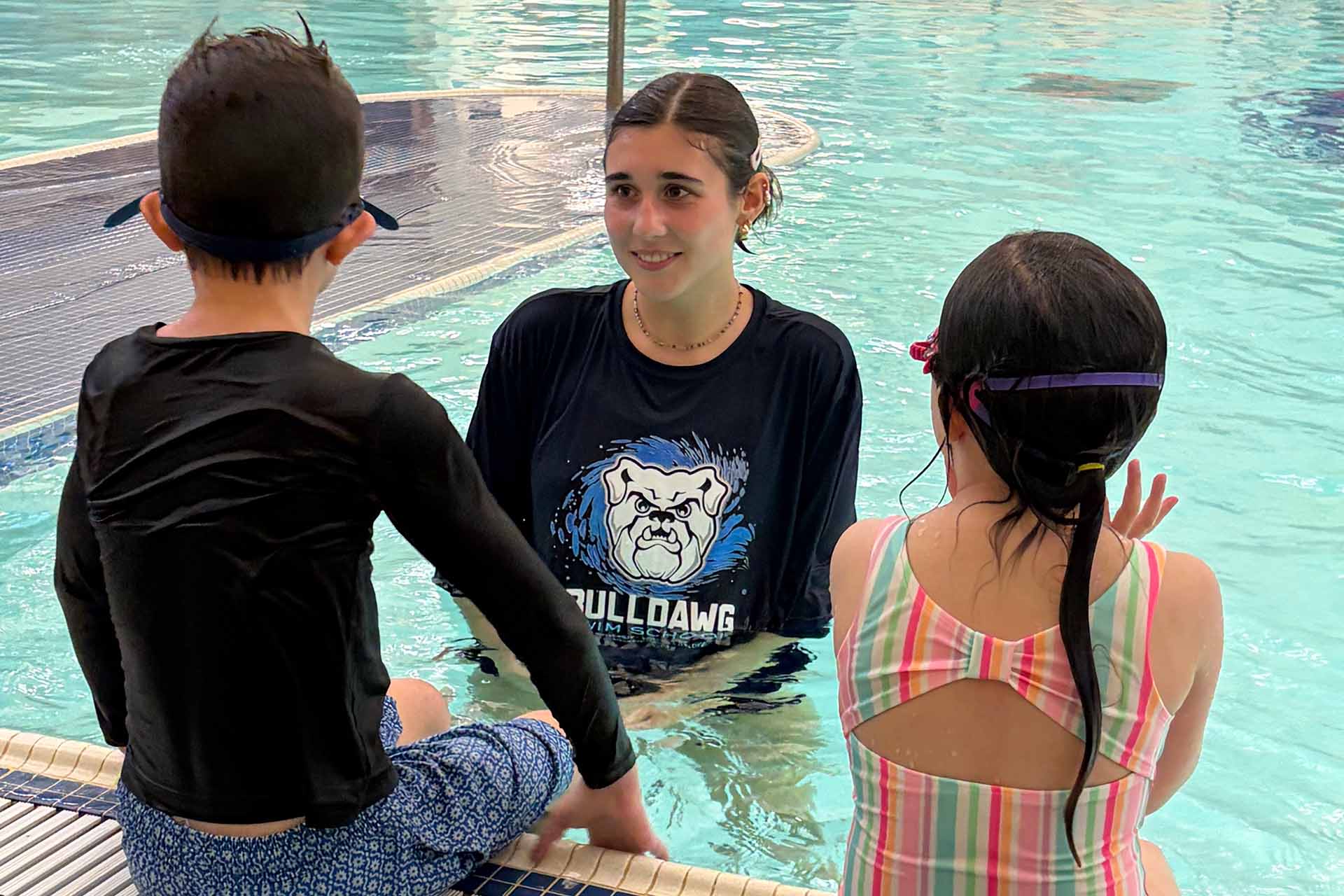 Female instructor Ali Quinn stands in pool speaking with child swimmers who are sitting on the edge of the pool.