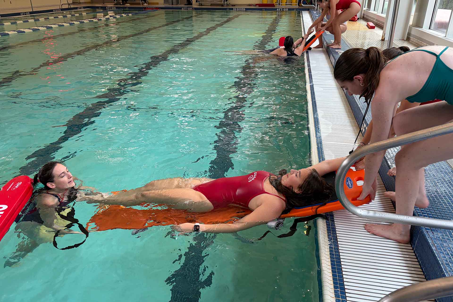 Two lifeguards lift a swimmer on a rescue board out of a swimming pool.