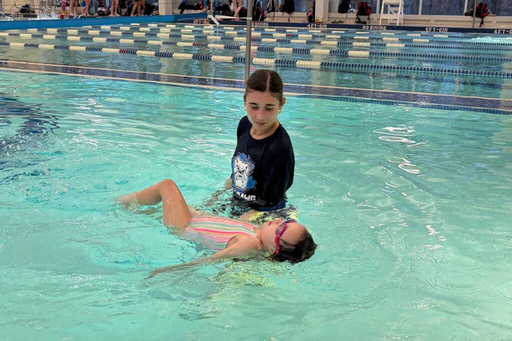 Female instructor stands beside child swimmer in pool during swimming lesson.