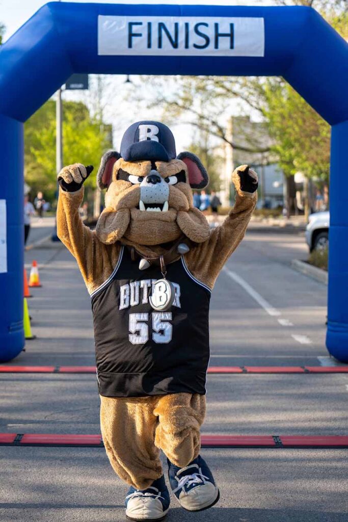 Hink the bulldog mascot raises his fists in the air while walking in front of a race finish line.