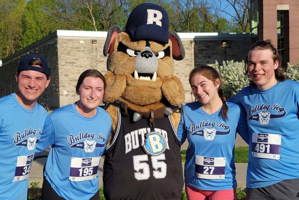 Group of young adults wearing matching blue Bulldog Jog shirts pose for a photo with Hink the bulldog mascot.