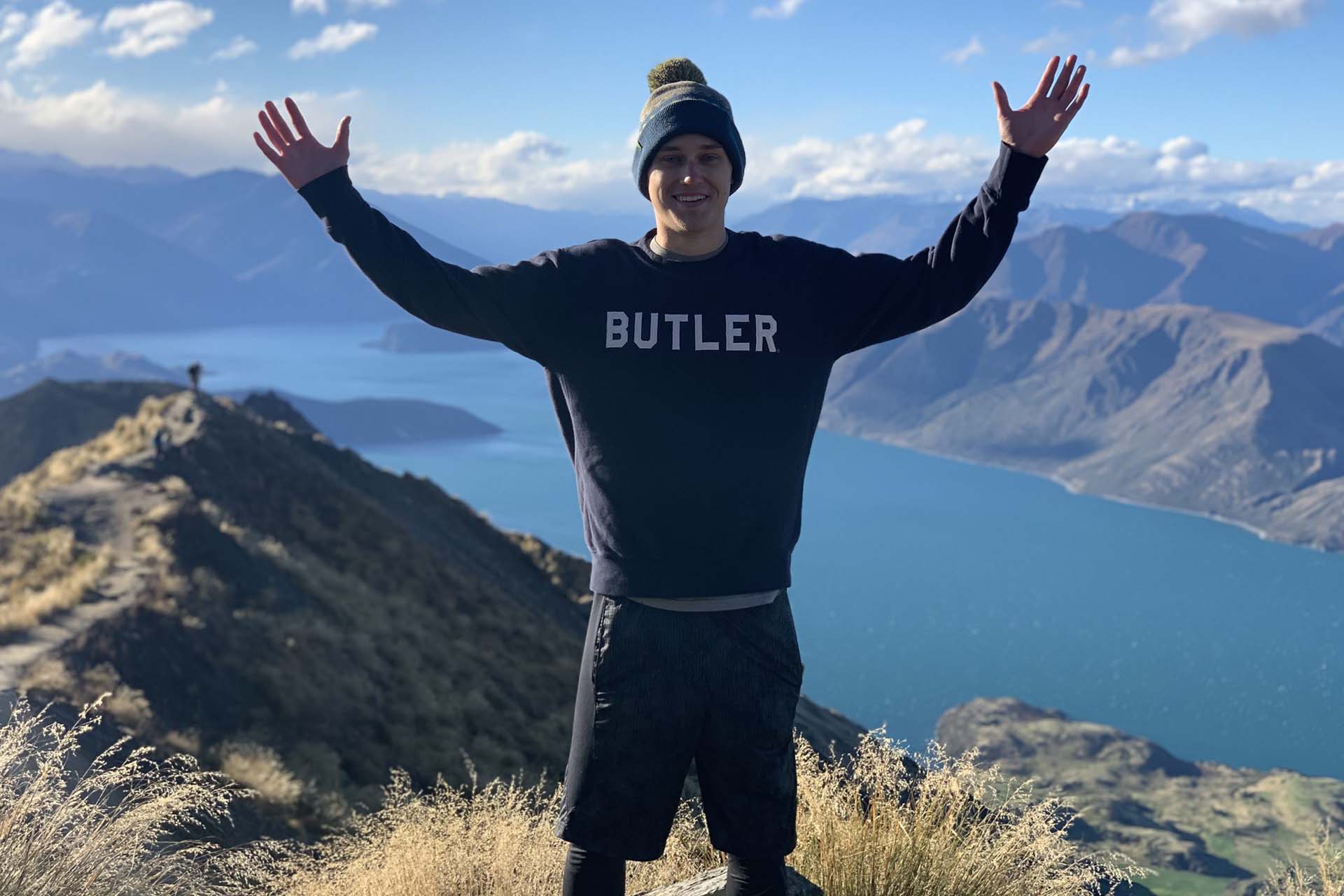 College student poses with arms in the air as he wears a Butler sweater and stands on a mountaintop overlooking a lake in New Zealand.