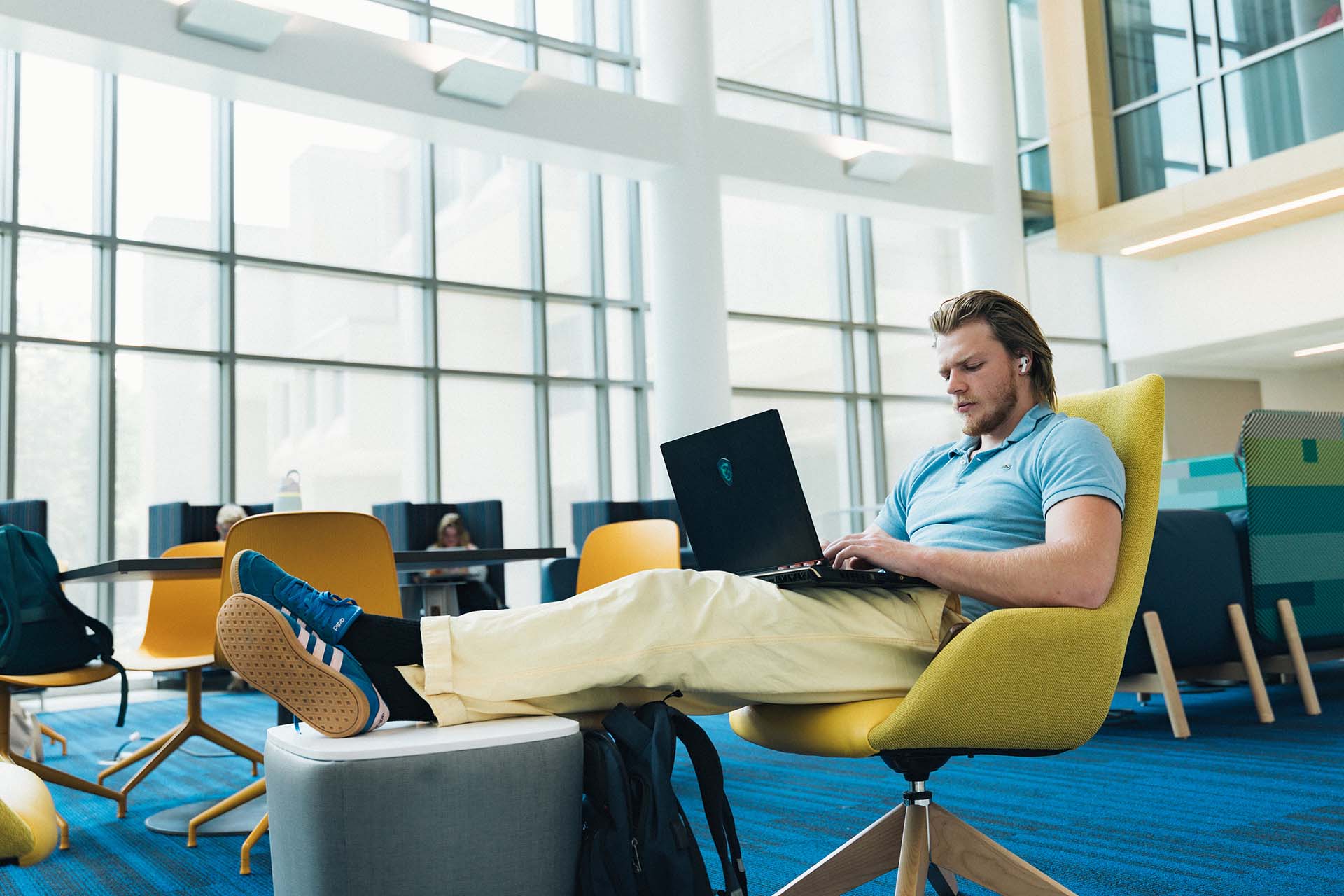Male college student reclines while working on laptop in bright atrium.