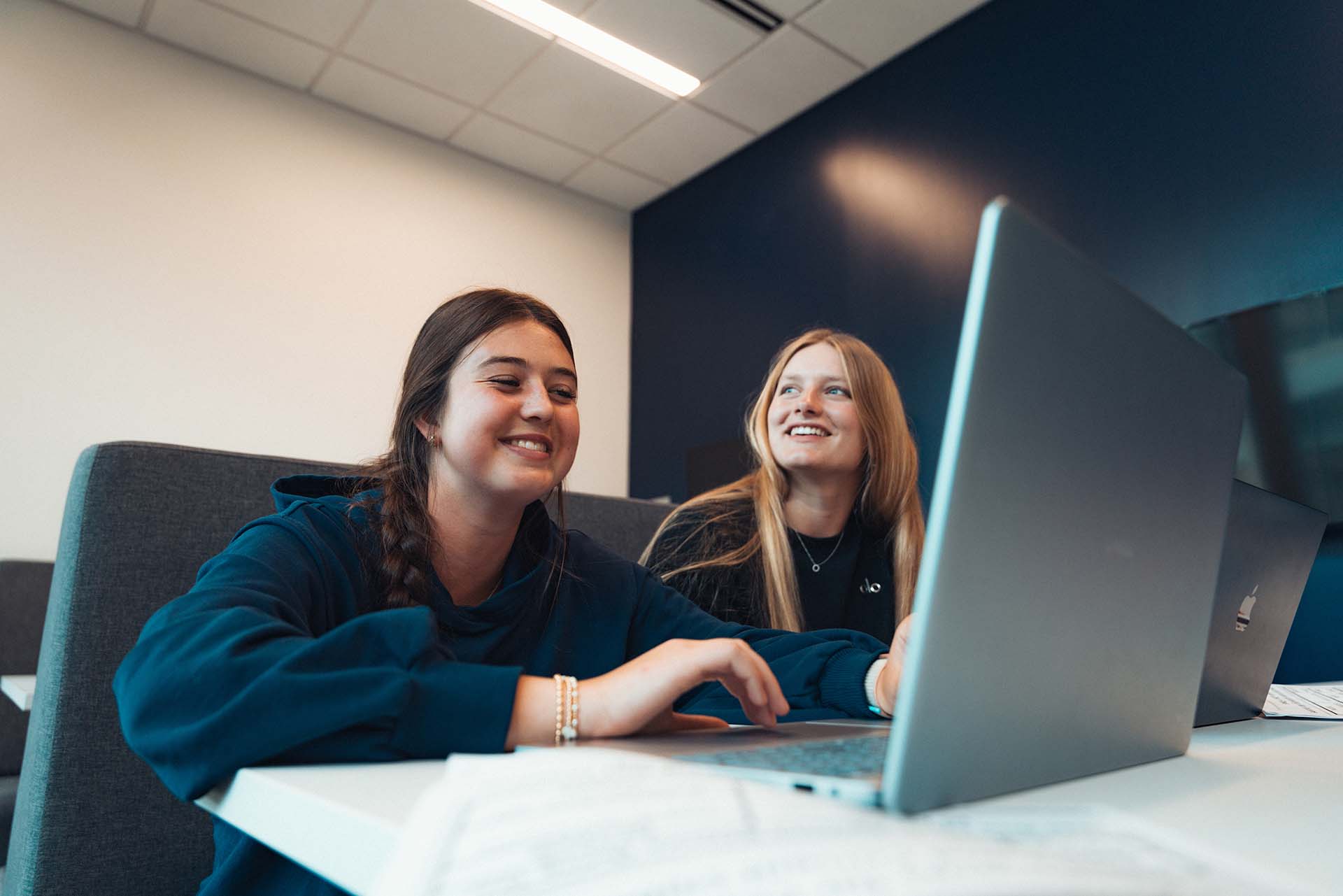 A pair of female college students smiles while working on a laptop together in a classroom.