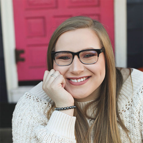 Mia Fazio, wearing eyeglasses and a sweater, leans on her wrist as she smiles brightly.