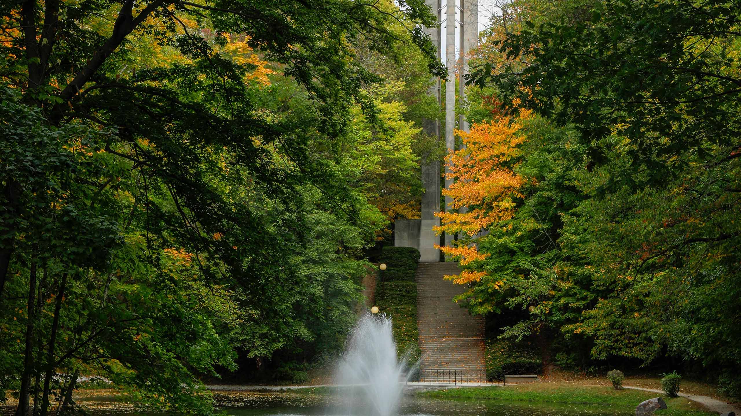Lush green trees with a twinge of orange leaves surround Holcomb Carillon tower and pond fountain.