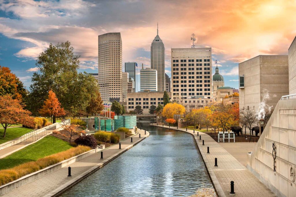 Downtown Indianapolis buildings, and canal with autumn foliage and concrete walkways alongside it.