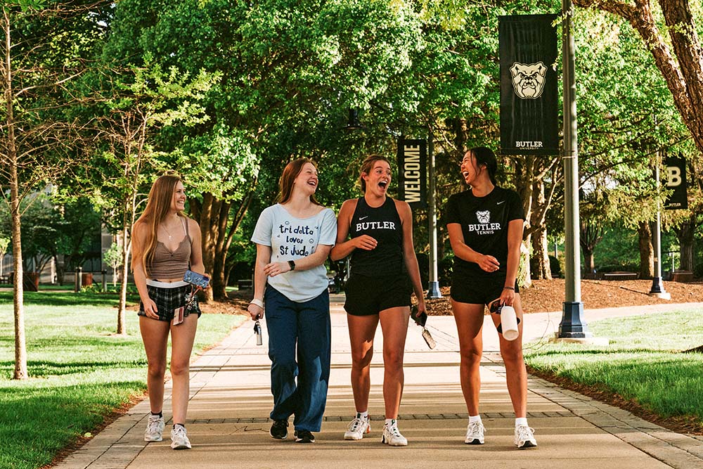 Group of four female friends walking down a campus path and laughing.