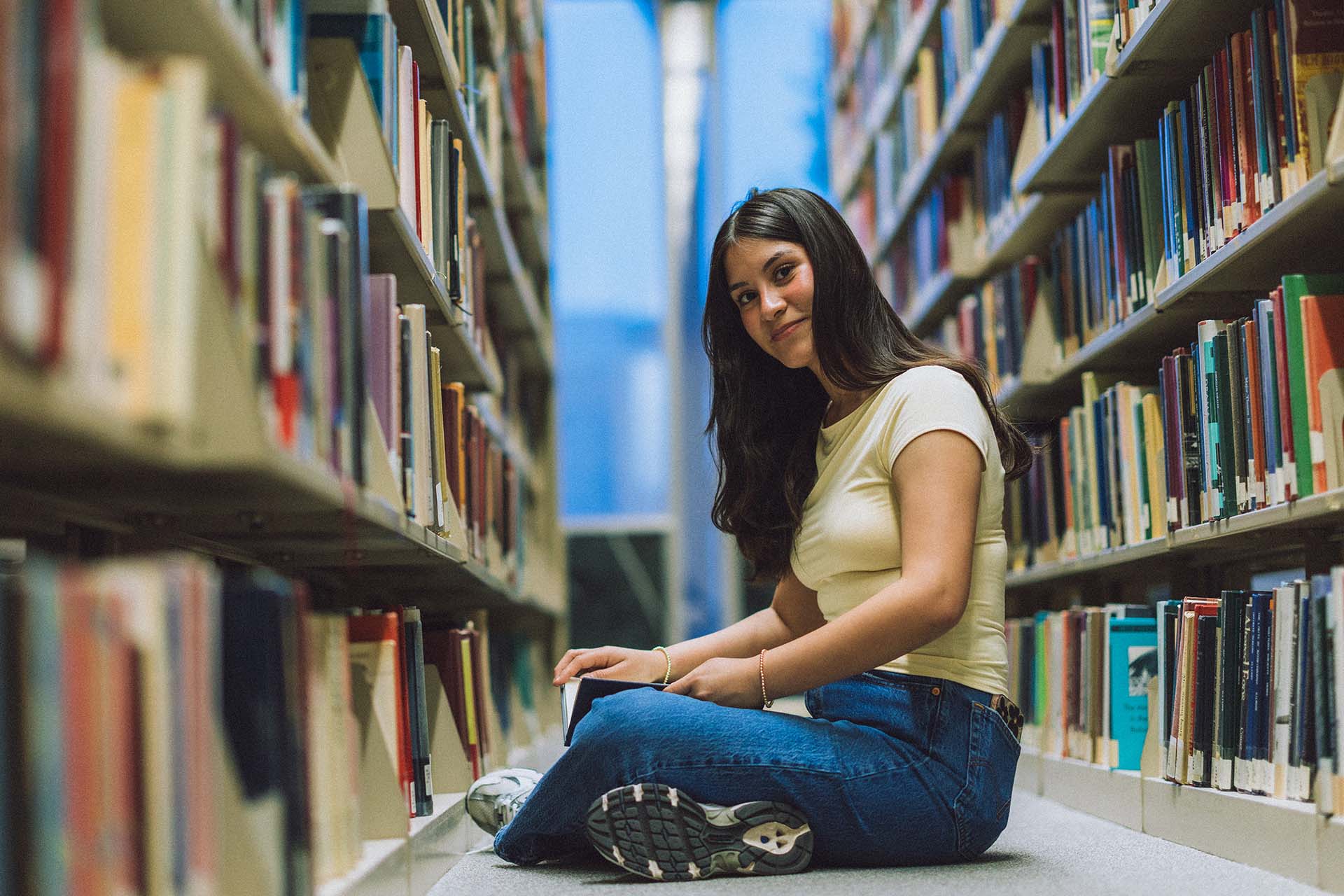 Portrait of college student sitting on floor in a library aisle surrounded by thousands of books.