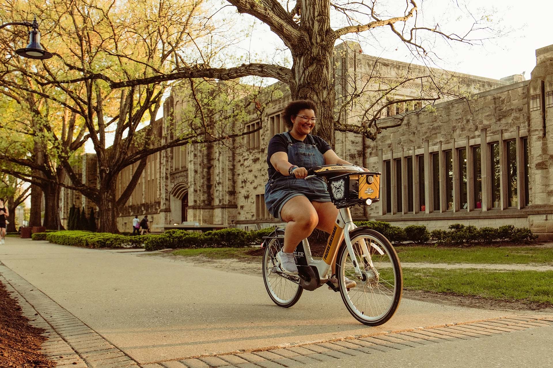 Woman riding a bicycle on a campus path with large limestone building and shade trees in background on a sunny day.
