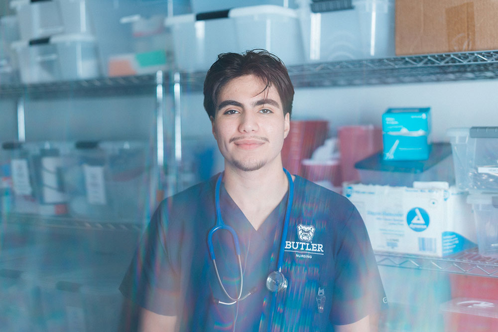 Male nursing student wearing scrubs poses for portrait in the supply room with shelves in the background and a flare effect.