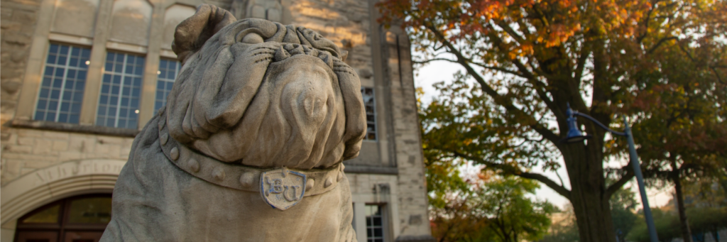 A stone statue of Butler Blue wearing a collar with the letters 'BU,' standing in front of Atherton Union, surrounded by autumn foliage.